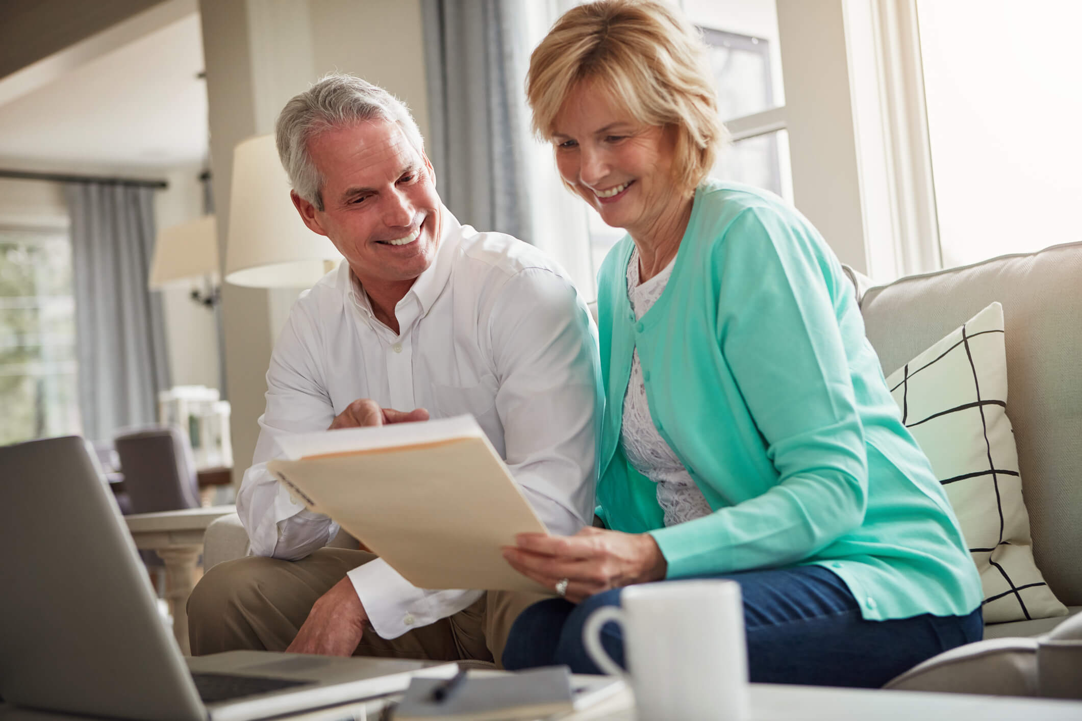 Senior couple, paperwork and laptop on sofa with discussion, smile and taxes in home. Old man, woman and computer with. Old man, women and computer with documents for compliance, reading and investment report with finance management.
